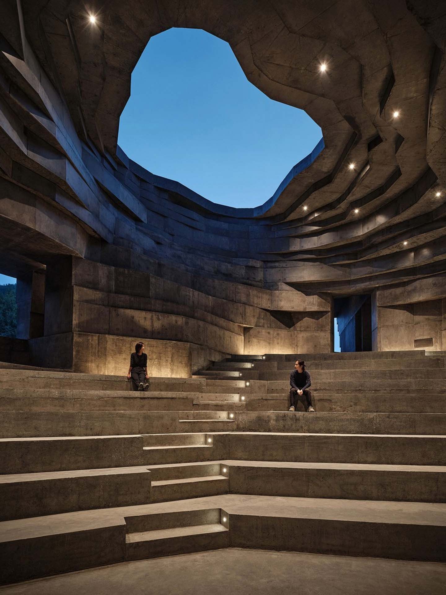 Two listeners on the amphitheater steps under the oculus at blue hour